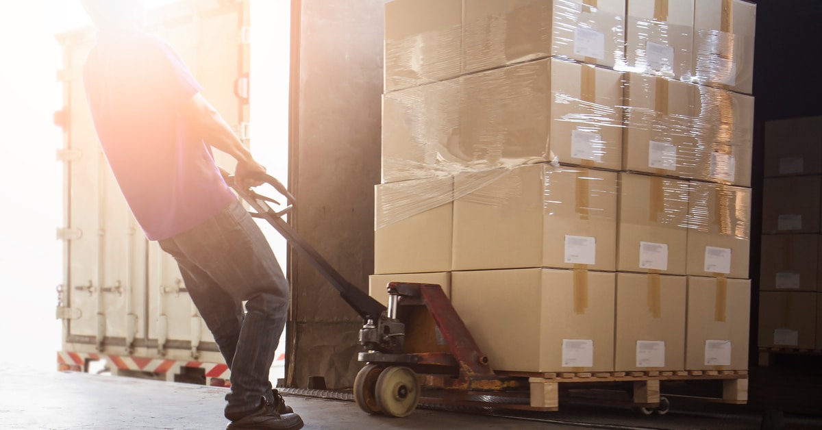 A warehouse worker unloads a pallet of stacked cardboard boxes from a semi-truck using a pallet jack with both hands.