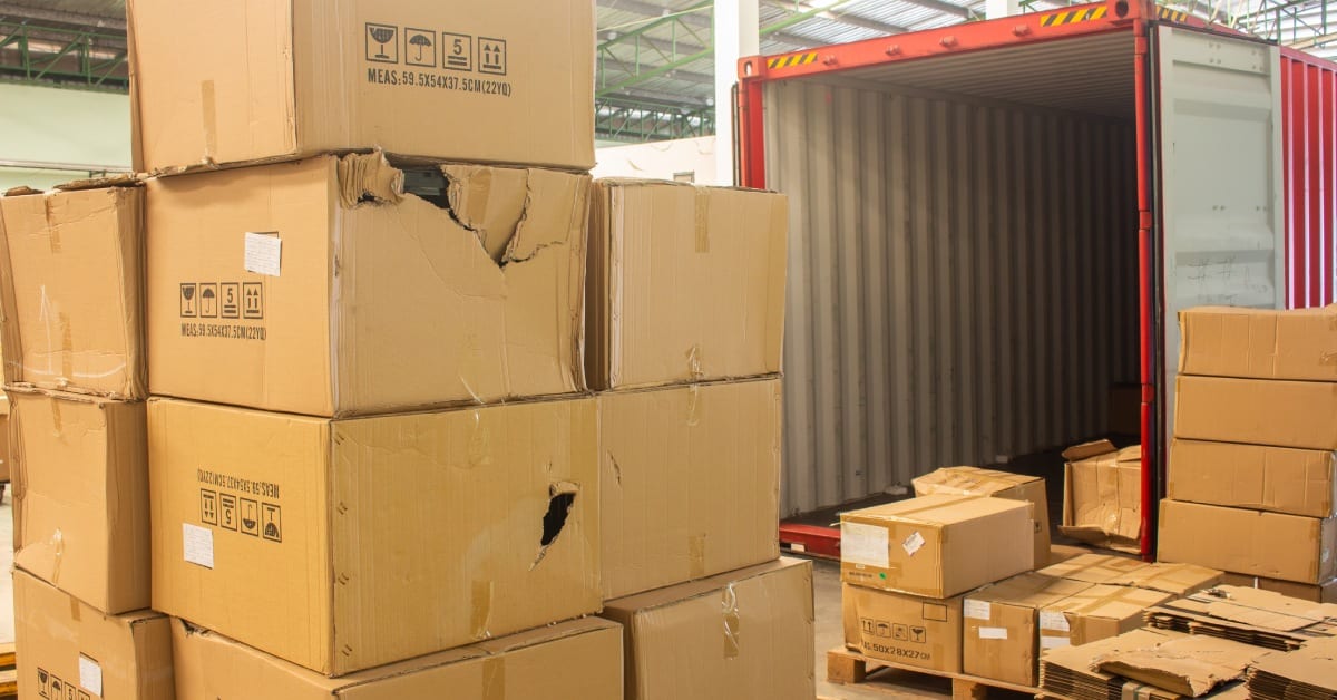 Stacks of unloaded, damaged cardboard boxes near an open red shipping container with cardboard boxes inside.