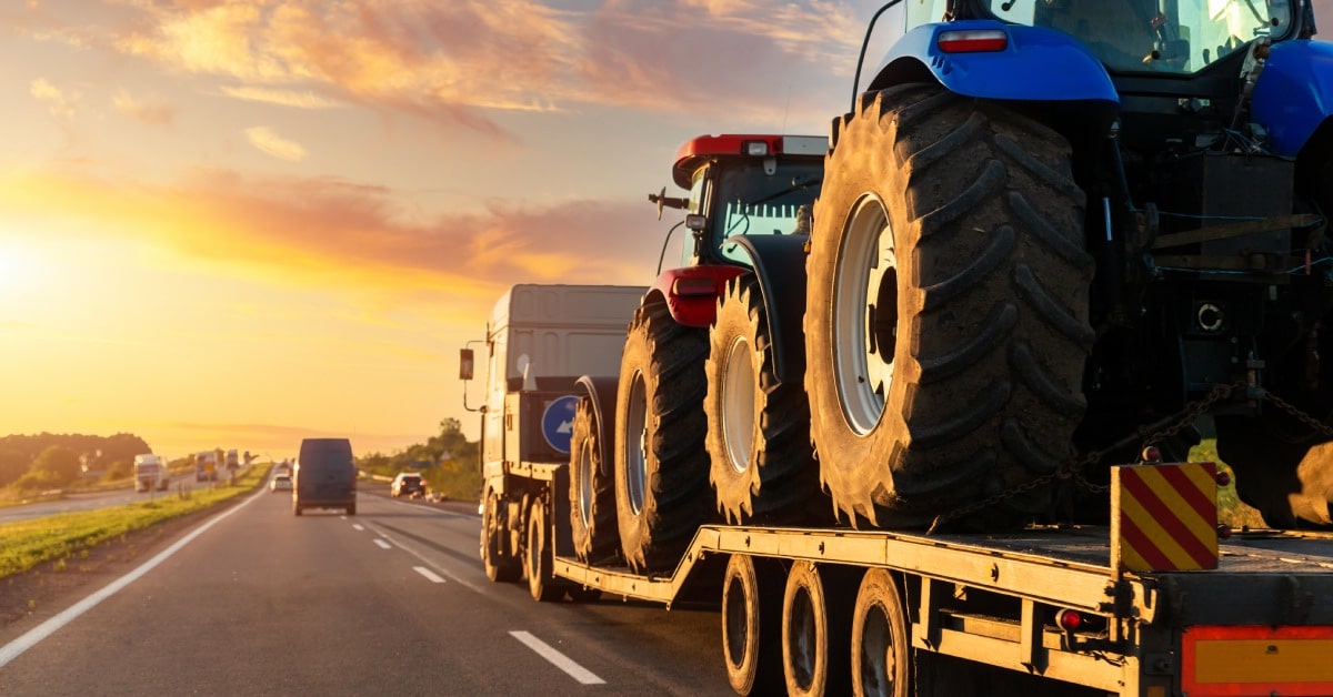 A heavy haul flatbed truck secured with a red and a blue farming trailer driving on a highway into the sunset.