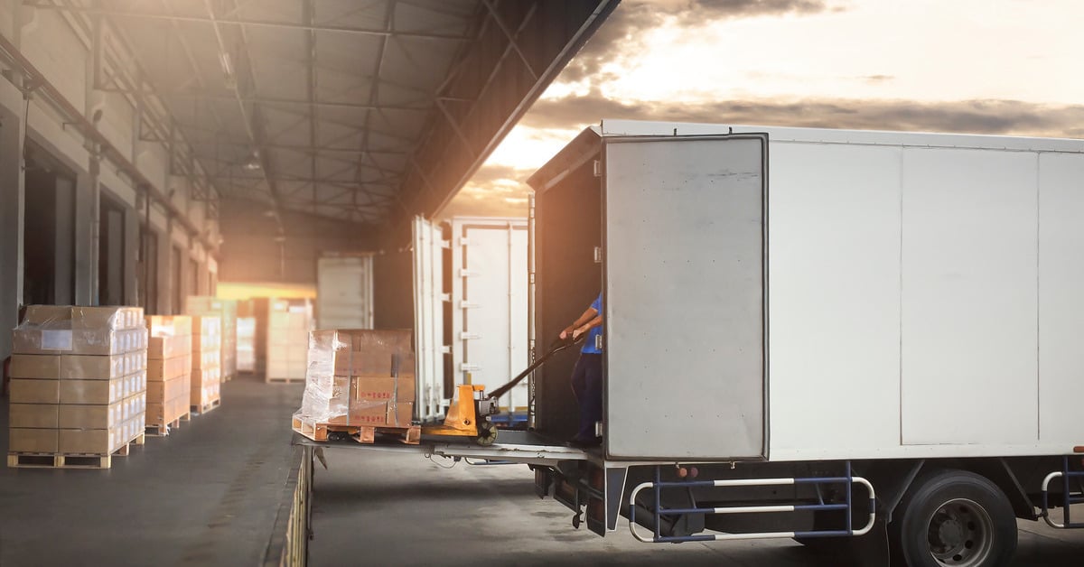 A man using a jack to load a cargo palate onto a white semi-truck trailer at a loading dock with other trailers.