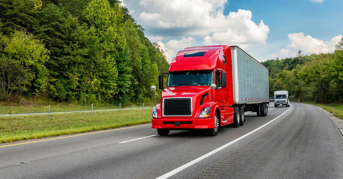 A red semi-truck with a metal trailer travels on a two-lane highway flanked by trees, with a van following behind.