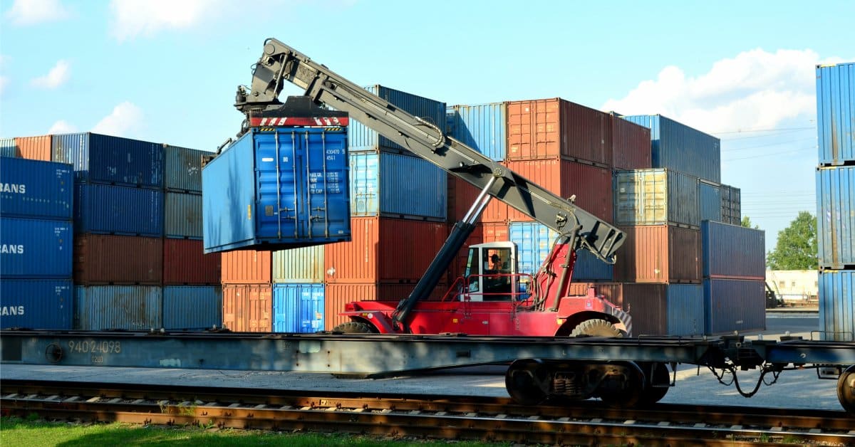 A red richtracker lifts a blue shipping container to place it on a freight rail system at a warehouse port.