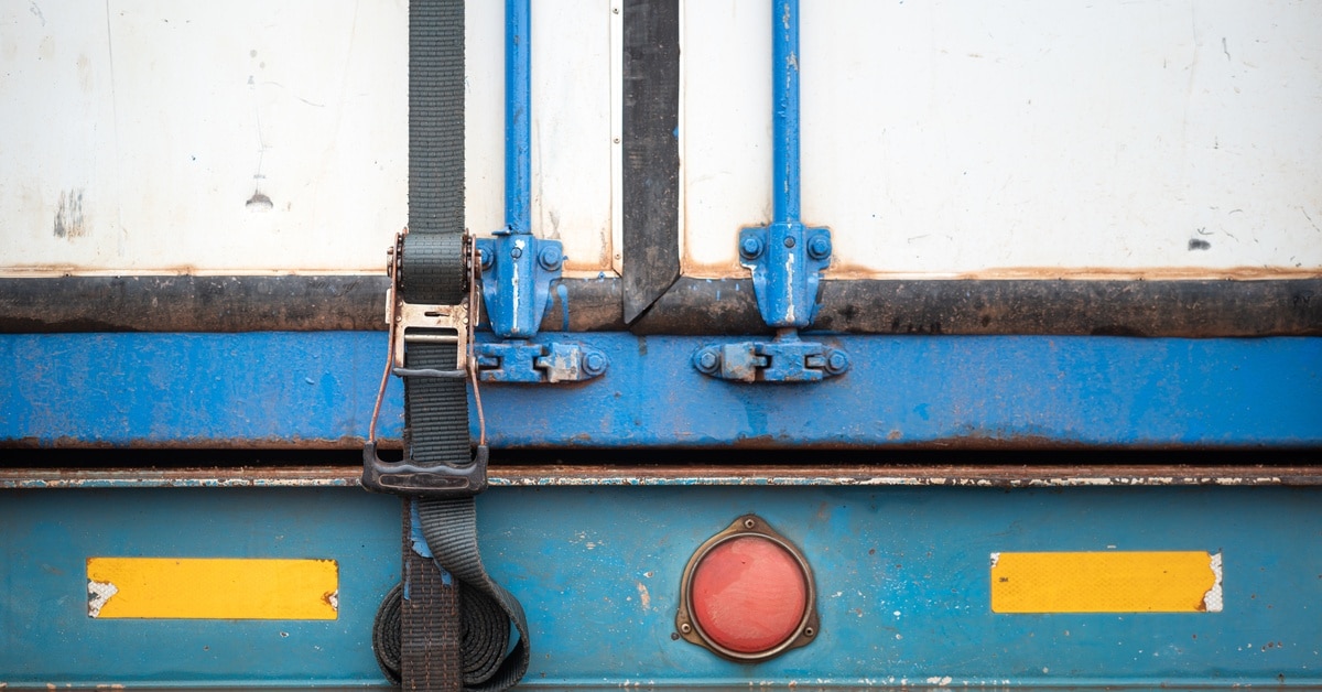 The back of a white cargo shipment trailer with a webbing belt strap that is latched down to secure the door.