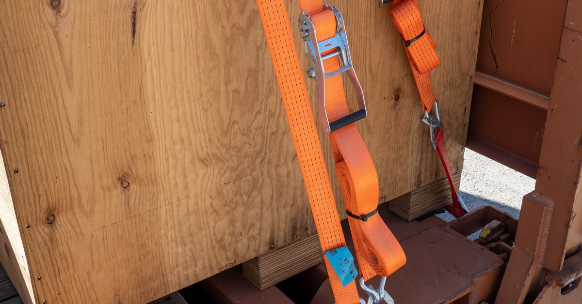 A close-up of a wooden cargo box loaded onto a pallet and secured by multiple orange ratchet straps.