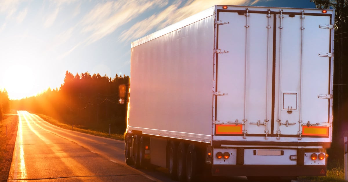 A white freight truck drives down a highway surrounded by trees. The back of the truck is visible.