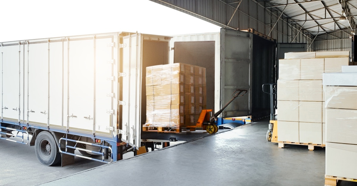 Multiple bulk pallets of cargo are loaded onto the back of a white shipping truck parked near a loading dock.