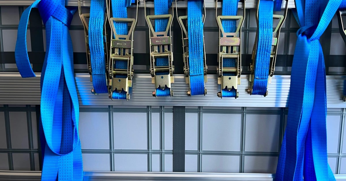 A group of multiple blue-colored ratchet tie down straps hanging next to one another. The straps have silver buckles.