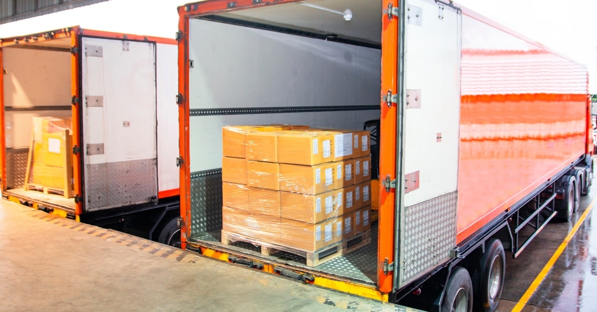 Two orange shipping trucks filled with pallets of cardboard boxes are parked at a warehouse loading dock.