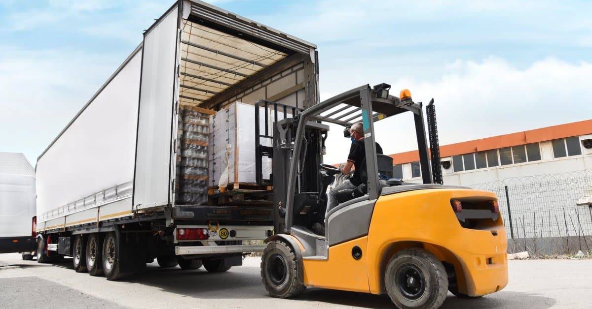 A yellow forklift is parked next to a large shipping truck while loading pallets of cargo. Load bars hold the cargo.