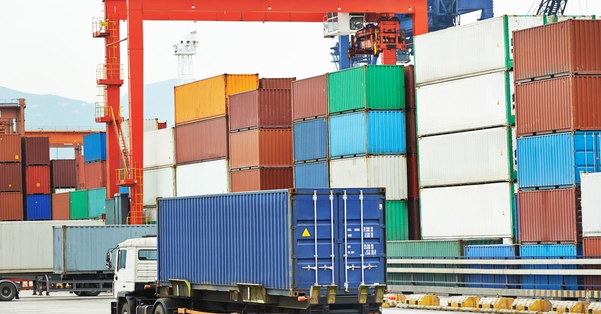 A freight truck carrying a blue shipping container drives around a cargo dock filled with multi-colored shipping containers.