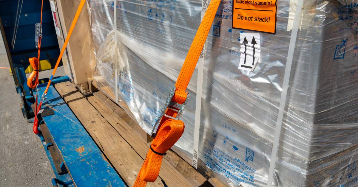 Multiple bright orange-colored ratchet straps secure wooden pallets of plastic wrapped cargo in place.