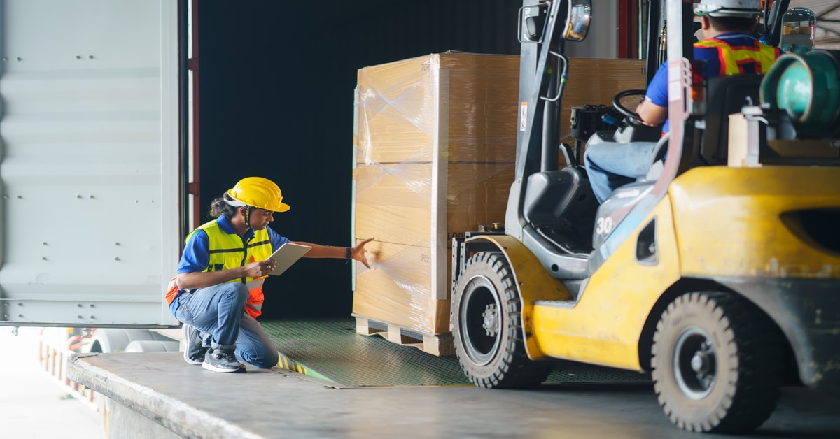 A worker wearing a protective hard hat kneels to check a pallet of cargo before it's loaded into a truck.