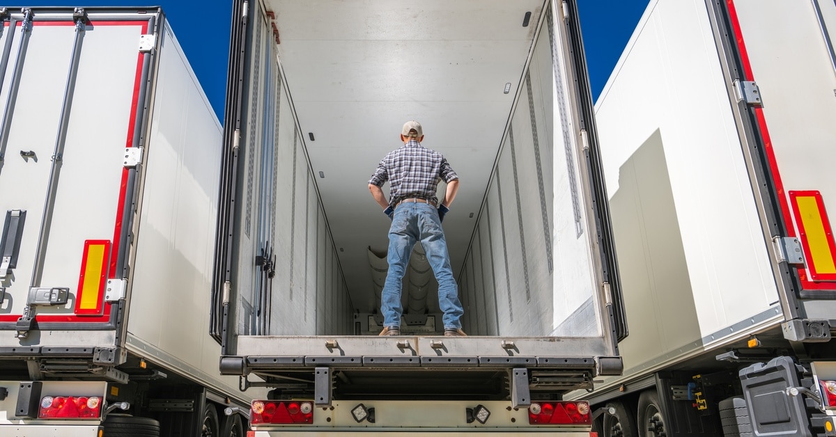 A man wearing a flannel shirt and jeans stands in the back of an empty semitrailer as he decides how to pack it properly.