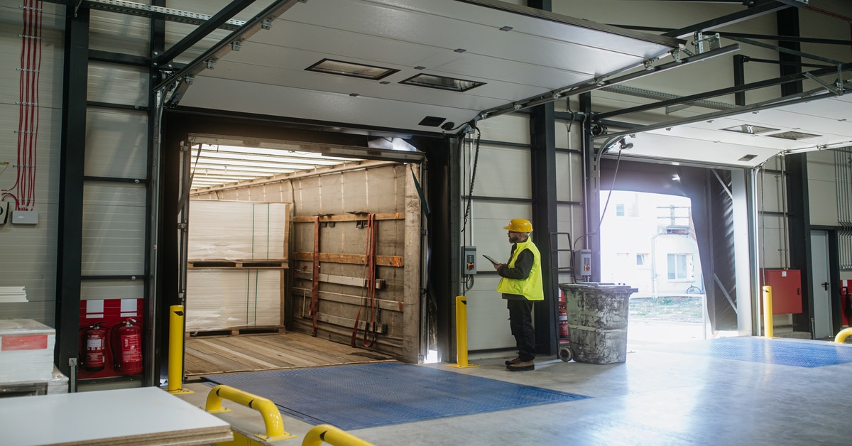 A worker wearing a yellow vest stands on a loading dock near an open door where a truck is being packed.