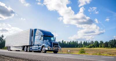 A blue semi-truck with a trailer driving on a rural road with a grassy field, forest, and blue sky in the background.